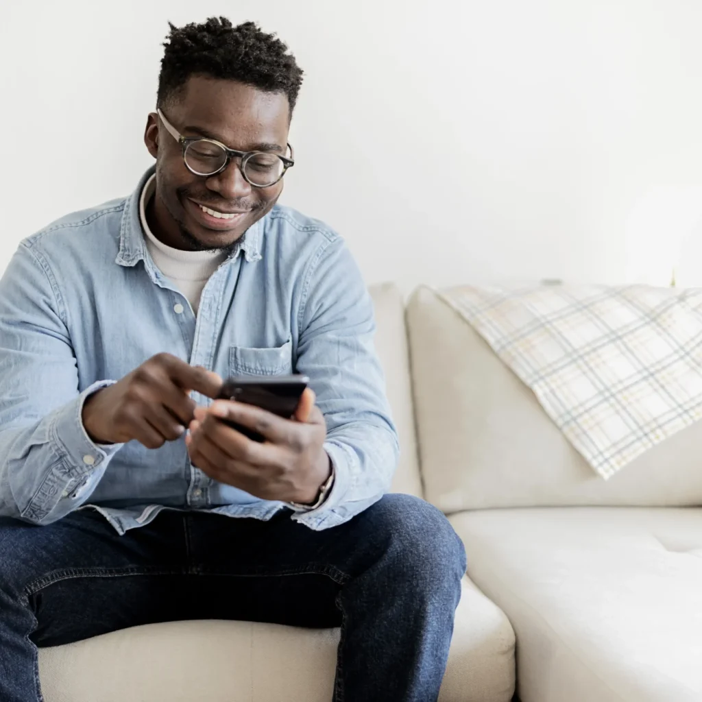 A man sits on a light-colored couch and smiling while using his smartphone.