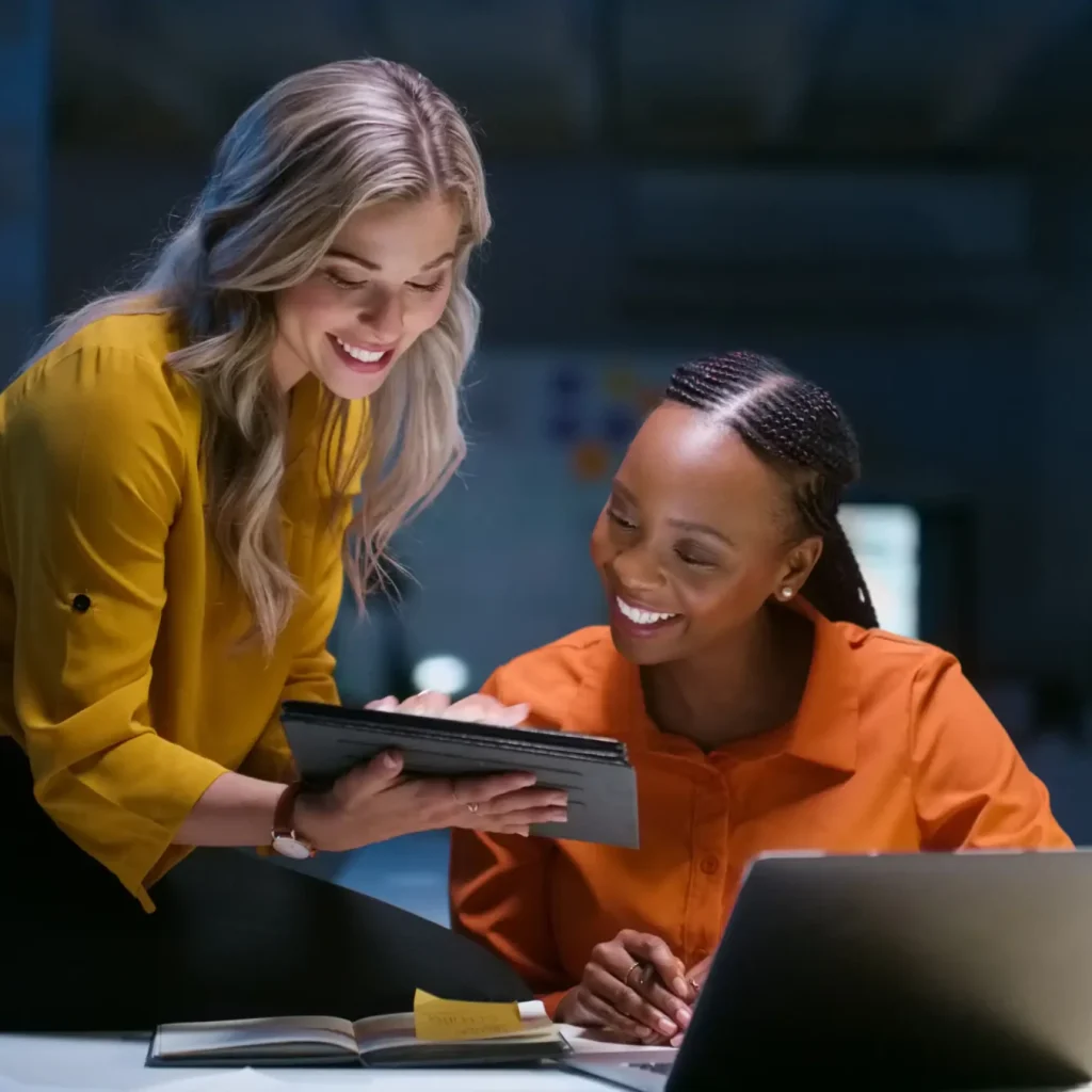 Two women are working together in an office. One woman is sitting at a desk with a laptop, while the other is looking at a tablet.